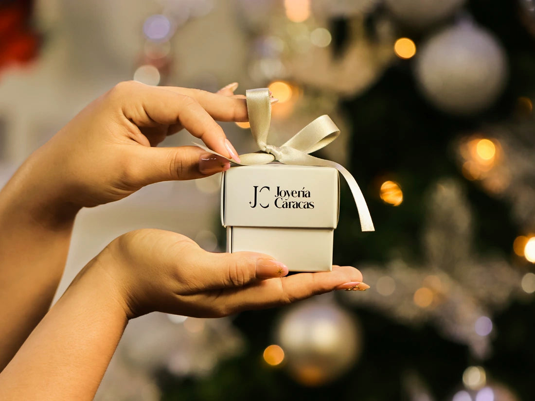 Manos de mujer sosteniendo una caja de regalo de Joyería Caracas frente a un árbol de navidad. 