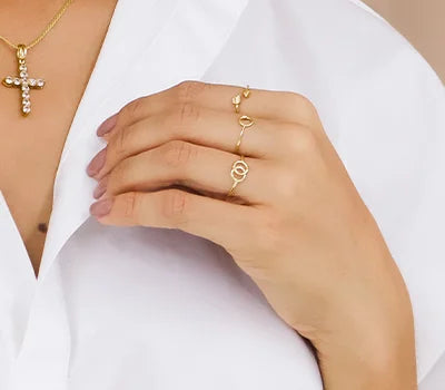 Mano tocando el cuello de una camisa blanca. La mano lleva tres anillos de oro: uno con un diseño simple y otros con un patrón intrincado,un nudo o una lazada. Además, la persona lleva un delicado collar de cadena de oro. Esta imagen podría ser relevante para discusiones sobre moda, diseño de joyería o cómo combinar accesorios con la ropa