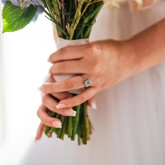 mano la cual sostiene un ramo de flores la cual lleva puesto un anillo en oro blanco de 18 quilates con una piedra de color celeste claro llamada topacio y alrededor de ella diamantes de color blanco hecho en Joyería Caracas.