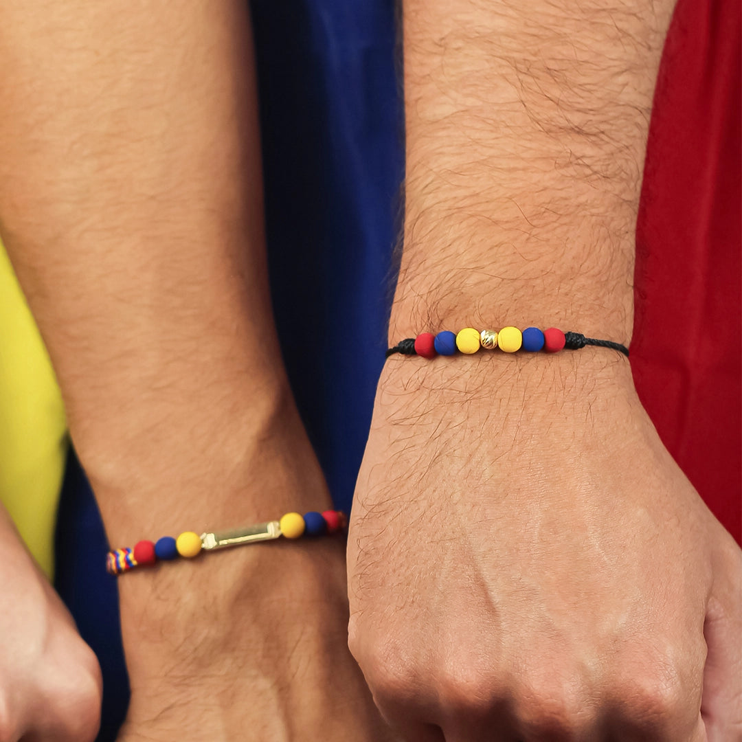 Mano empuñada mirando hacia abajo con pulsera tejida conformada por hilo de color negro, pepas de color amarillo, azul y rojo y esfera con textura de oro de color amarillo de 18 quilates. 