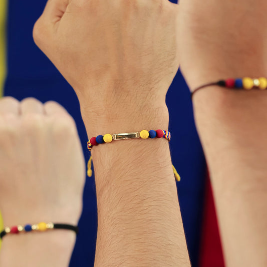 Mano empuñada hacia arriba con pulsera tejida la cual esta conformada por hilo de color amarillo, azul y rojo, pepas con los mismos colores y placa lisa en oro de color amarillo de 18 quilates.