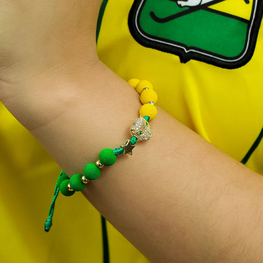 Mano de mujer con pulsera tejida, la cual esta conformada por hilo de color verde, pepas de color verde y amarillo, esferas lisas, dije en forma de estrella y en el centro dije de un leopardo en oro amarillo de 8 quilates, el cual esta conformado por cristales de color verde en los ojos y en el alrededor cristales de color blanco. 