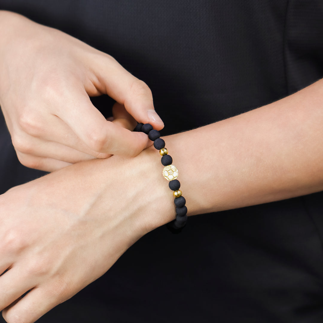 Mano de hombre con pulsera tejida en hilo negro con esferas lisas y dije en forma de balón en oro amarillo de 18 quilates, hecho en Joyería Caracas. 