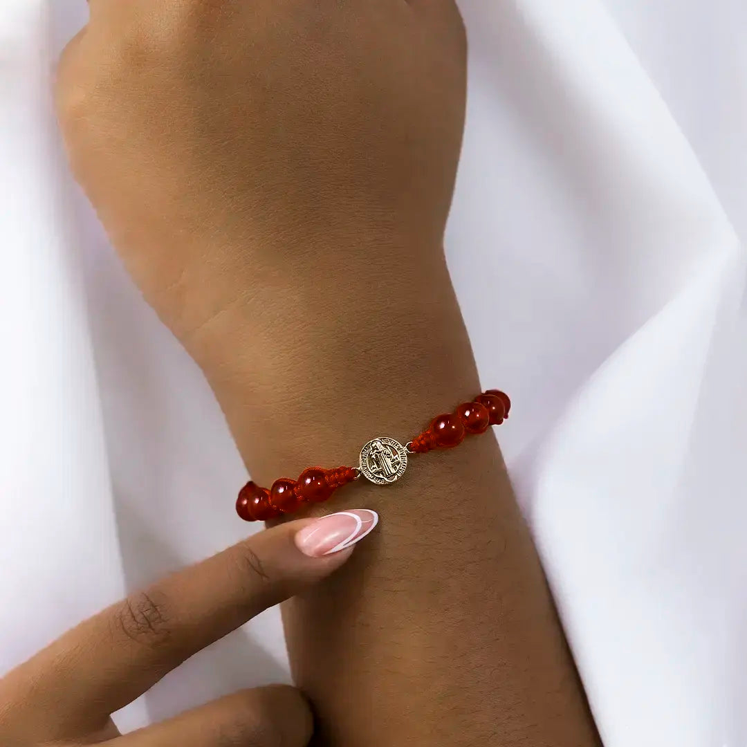 Mano de mujer con pulsera tejida en hilo rojo, piedras rojas y dije de San Benito en oro amarillo de 18 quilates, hecho en Joyería Caracas. 