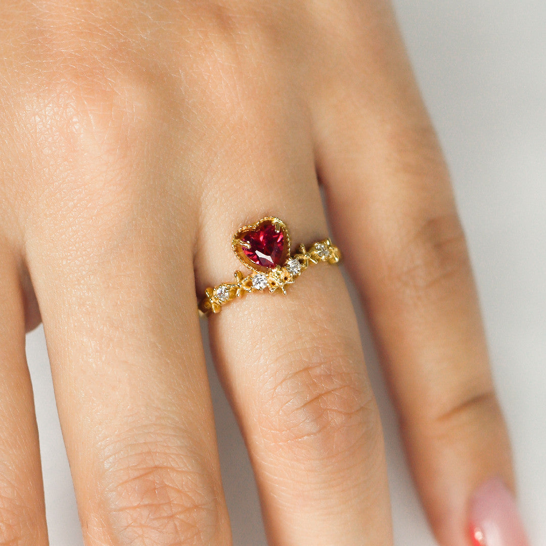 Mano con anillo para mujer, hecho en oro de 18k, color amarillo, el cual tiene una piedra de color rojo en forma de corazón y cristales debajo de ella, ubicado en el dedo anular.