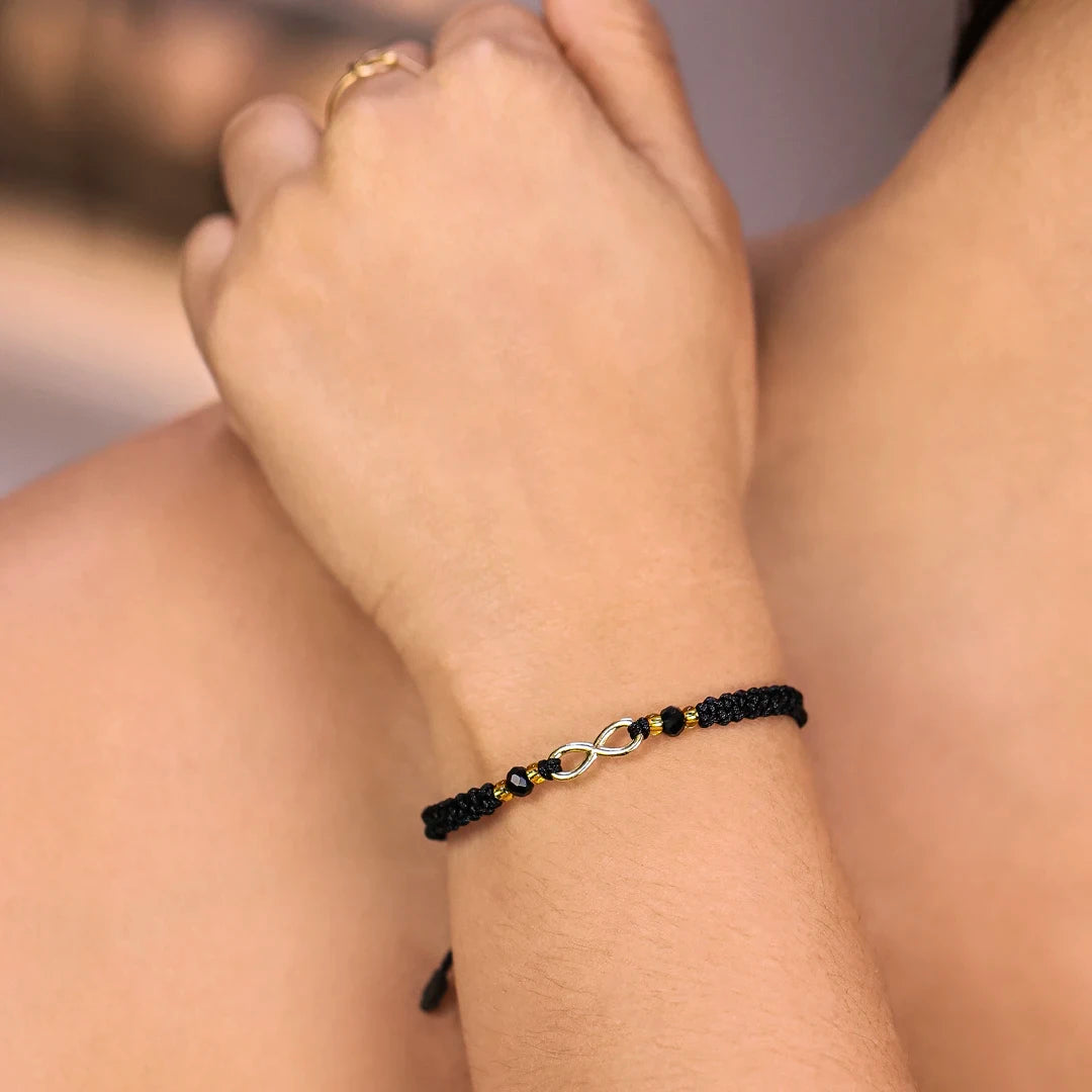 Mano de mujer con pulsera tejida en hilo negro coreano, accesorios de bisutería en color negro, dorado y dije en forma de infinito en oro amarillo de 18 quilates, hecho en Joyería Caracas. 